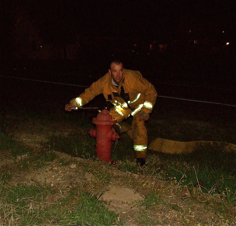 Image: Here comes the water! — Without a moment to lose, Italy Firefighter, Paul Cockerham, connects the hose and rushes water to Italy’s Engine 1 truck. With thousands of gallons of water needed to douse the fire raging inside Milford Cash Grocery, the town’s water tower was nearly emptied.