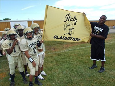 Image: See us now? — IYAA A-Team Gladiators’ Coach Edwin Wallace and the players hold up the ‘Italy Gladiators’ flag before taking the field.