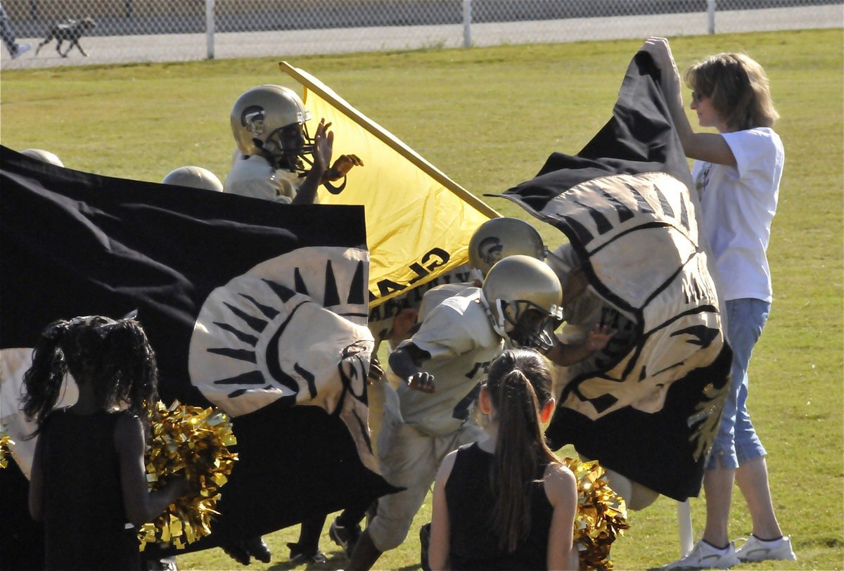 Image: Dramatic entrance — The IYAA B-Team Gladiators bust through the banner while carrying the school colors and being cheered on by the IYAA Minors Cheerleaders.