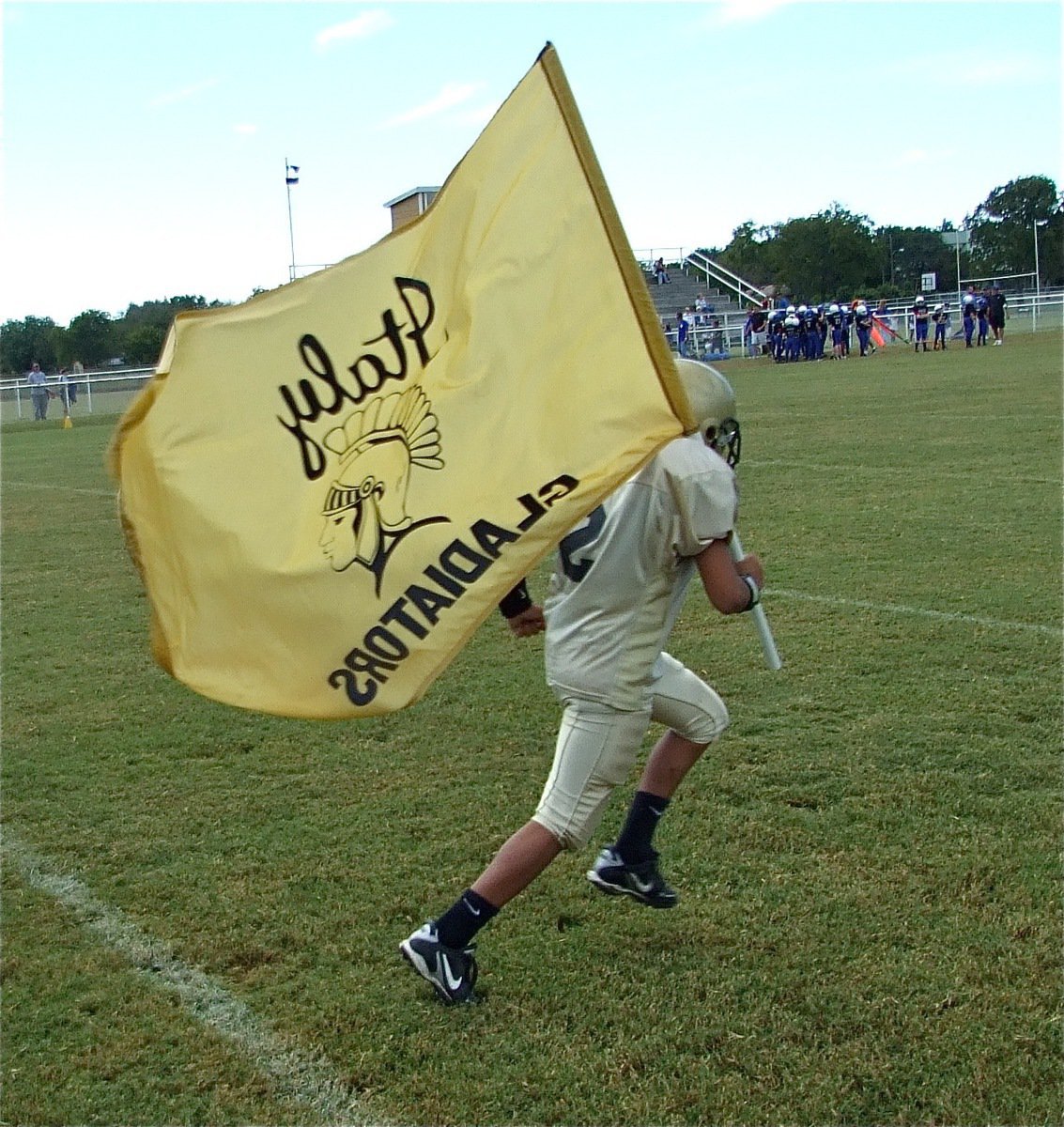 Image: Tylan Wallace — IYAA A-Team quarterback Tylan Wallace leads his teammates onto the field while hoisting the ‘Italy Gladiators’ flag.