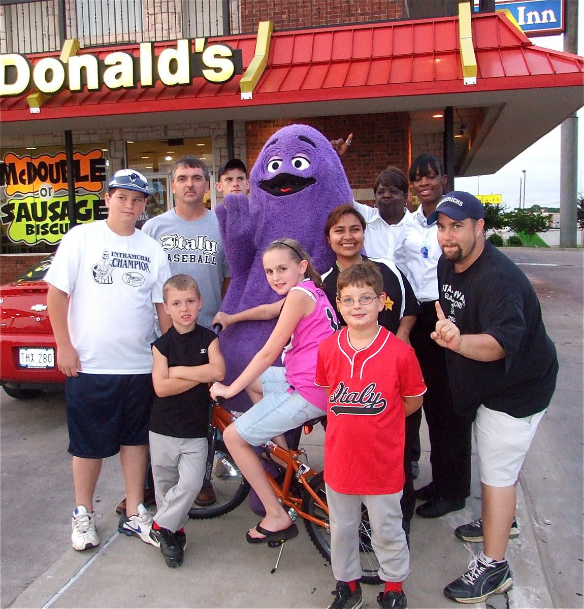 Image: IYAA players and coaches celebrate with McDonald’s® staff — Kirby Nelson wins the bicycle raffle during IYAA Sports Night at McDonald’s in Italy. Back row: IYAA president Gary Wood, Justin Wood, Grimace™ (Keanan Brown), Brenda Smith and Jakiesha Williams. Middle row: John Byers, Bryce DeBorde, Bike raffle winner Kirby Nelson, Maria Balderas and IYAA vice president Barry Byers. Front: Gage Wafer.