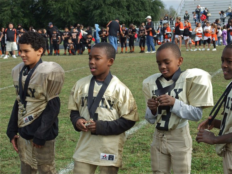 Image: IYAA B-Team Gladiators receive their Superbowl Champion medals — Tylan Wallace(7), Kendrick Norwood(1), Addison Alexander(3) and Jaylon Davis(4) stand proud as they pose with their Superbowl Champion medals during the awards ceremony after the game. B-Team, head coached by Ken Norwood, defeated the Ferris Yellowjackets 19-0.