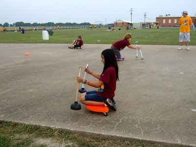 Image: These third graders are participating in the scooter plunger race. The students have to push themselves on the scooters by using the plungers and get to the finish line first to win.