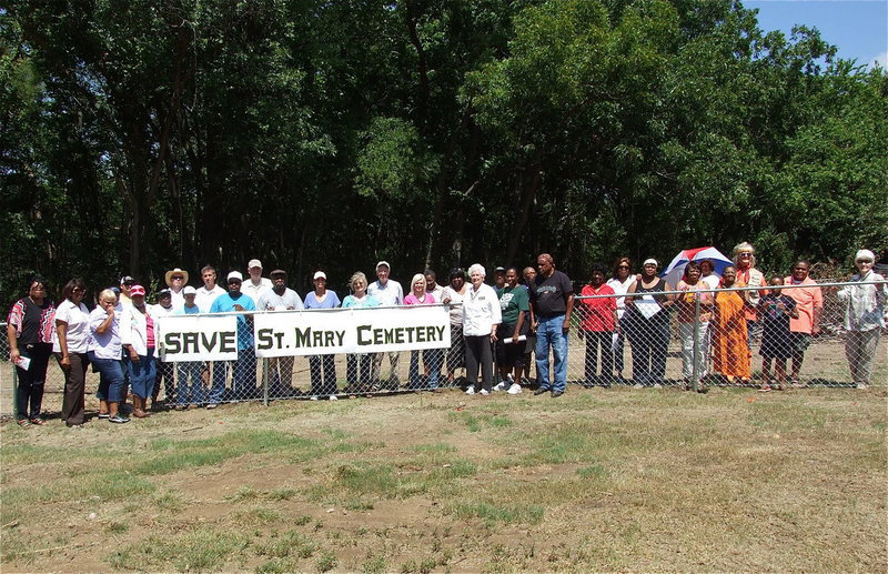 Image: Activists and guests who attended the SAVE St. Mary Cemetery rally in Ellis County, Texas pose around a sign hung by the Elmerine Bell Family and supporters on the fence that was added by Creek Land &amp; Cattle Company after the land developer destroyed a preexisting fence line while creating a road thru a portion of St. Mary Cemetery, destroying gravestones in the process that dated back to the 1870s.