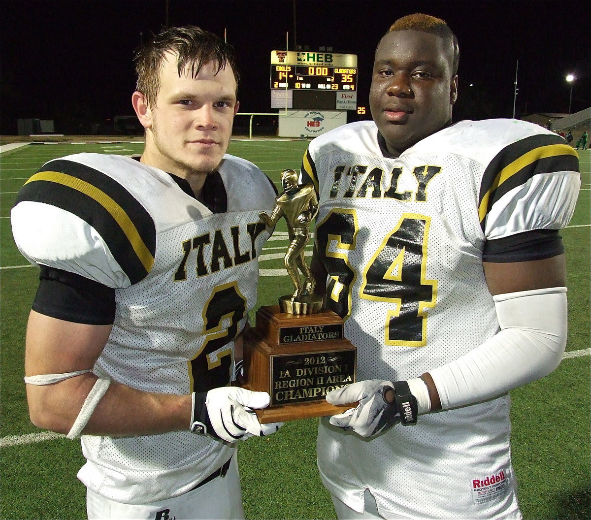 Image: Proud seniors Chase Hamilton(2) and Adrian Reed(64) with the area championship trophy as the final score reflects off the sculpture, Gladiators 35 and Eagles 14.