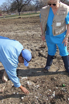 Image: While Elmerine Bell sorts thru the scattered debris, Deborah Franklin, aka Dr. Graveyard (on the right), comments, “This is not a landfill we are standing in. These glass and bottle fragments are here because they decorated the headstones that marked the graves.”