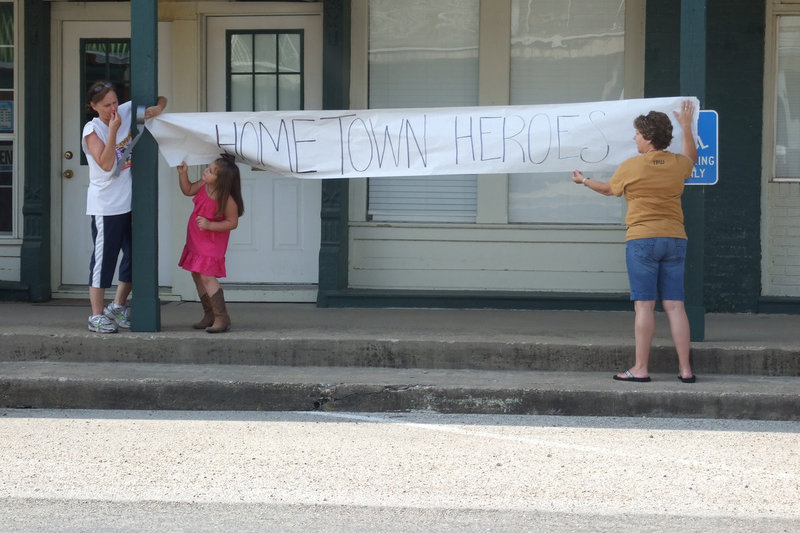 Image: With little Tinzley’s help, Sandy Eubank and Dian Morrison hang a banner that reads, “Hometown Heroes,” in anticipation of Italy High School’s student-athletes about to be honored during a well-deserved parade to recognize their recent accomplishments in sports.