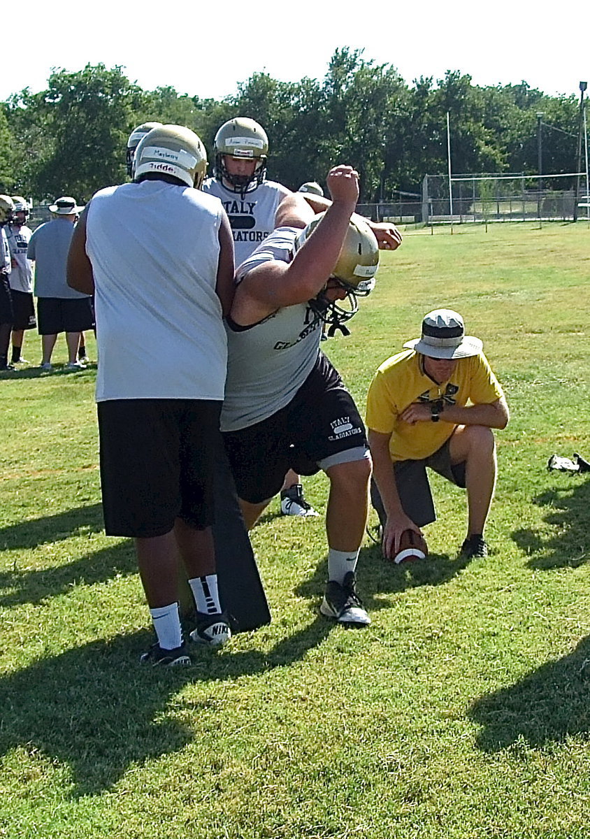 Image: With coach Jon Cady simulating the snap, senior Gladiator Kevin Roldan demonstrates the rip technique during a defensive line drill with teammate Darol Mayberry offering resistance.